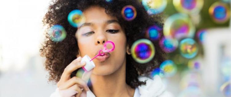 Photo of a woman holding a pink bottle of bubble solution while blowing into a bubble wand. Bubbles are floating off into the air.