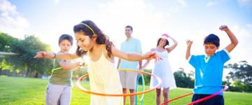 Photo of a mom, dad, and three kids in a park on a sunny day. The kids are playing with hula hoops.