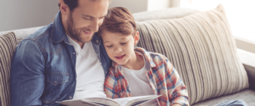 Photo of a man and his young son reading a book together on a sofa