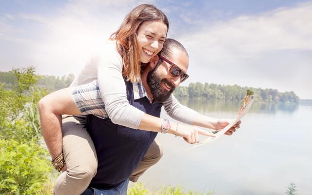 Photo of a smiling couple by a lake, a man holds a woman on his back while she holds out a map for them both to see