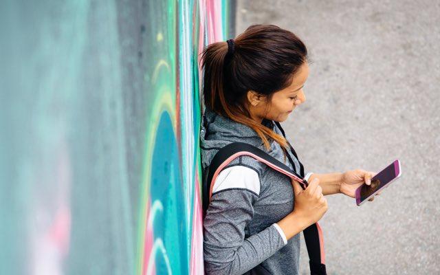 Photo of a woman leaning against a wall decorated with graffiti. She is looking at her phone.