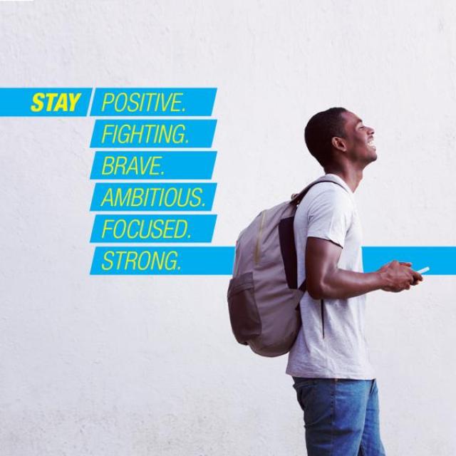 A young man wearing a backpack laughs while looking at his phone. The text behind him reads "Stay positive. Fighting. Brave. Ambitious. Focused. Strong."